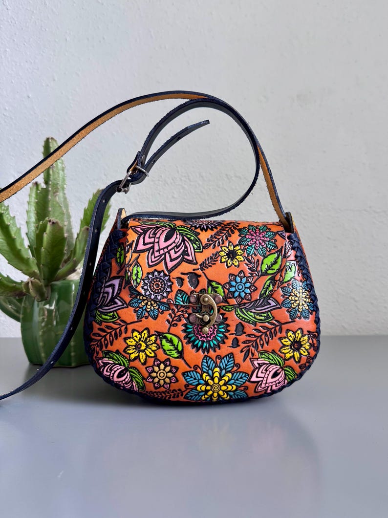 May include: A leather crossbody bag in orange, decorated with a floral pattern in pink, blue, yellow, and green. It features a dark blue strap and trim, and a metal clasp. A cactus plant is in the background.