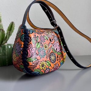 May include: An orange leather handbag with a vibrant floral design. The bag has a black strap and handle, and a blue braided detail. The bag is approximately 25 cm wide. A stylish accessory.