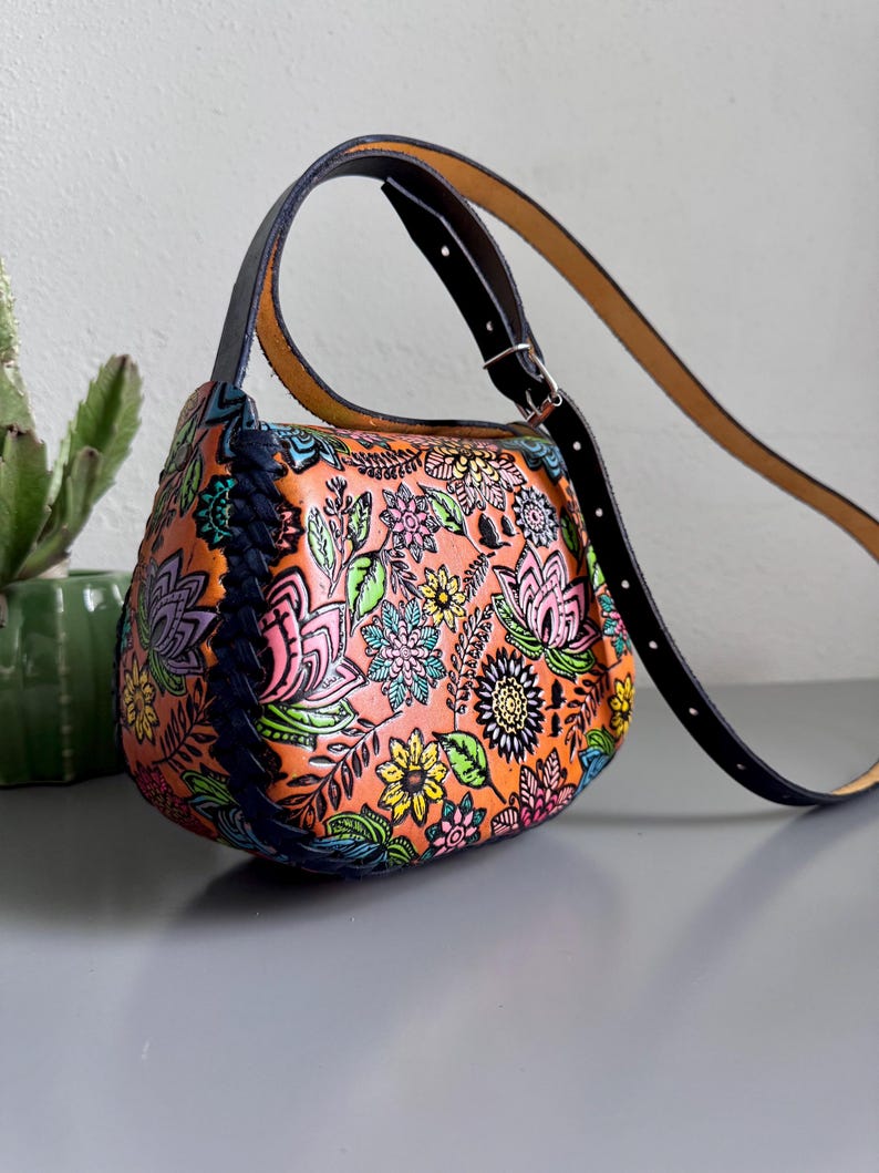 May include: An orange leather handbag with a vibrant floral design. The bag has a black strap and handle, and a blue braided detail. The bag is approximately 25 cm wide. A stylish accessory.