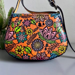 May include: Orange leather crossbody bag with a colourful floral design. The bag features a black strap and a flap closure. The flowers are pink, yellow, blue, and green, set against an orange background.