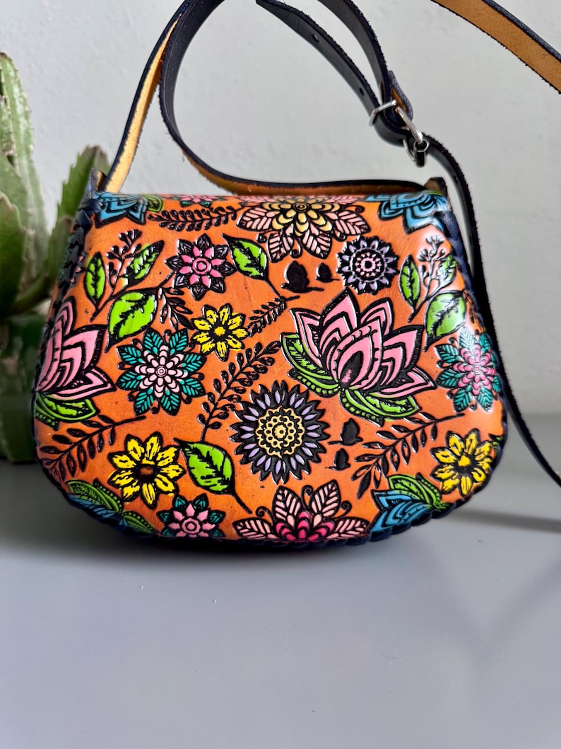 May include: Orange leather crossbody bag with a colourful floral design. The bag features a black strap and a flap closure. The flowers are pink, yellow, blue, and green, set against an orange background.