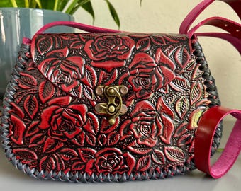 May include: A small, red leather crossbody bag with a floral rose pattern. The bag features a brass clasp, a pink strap, and a gray stitched border. The roses are embossed on the leather.