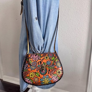 May include: Orange leather crossbody bag with a floral design in pink, green, blue, and yellow. The bag has a brown strap and a metal clasp. The bag is approximately 23 cm wide and 18 cm tall.