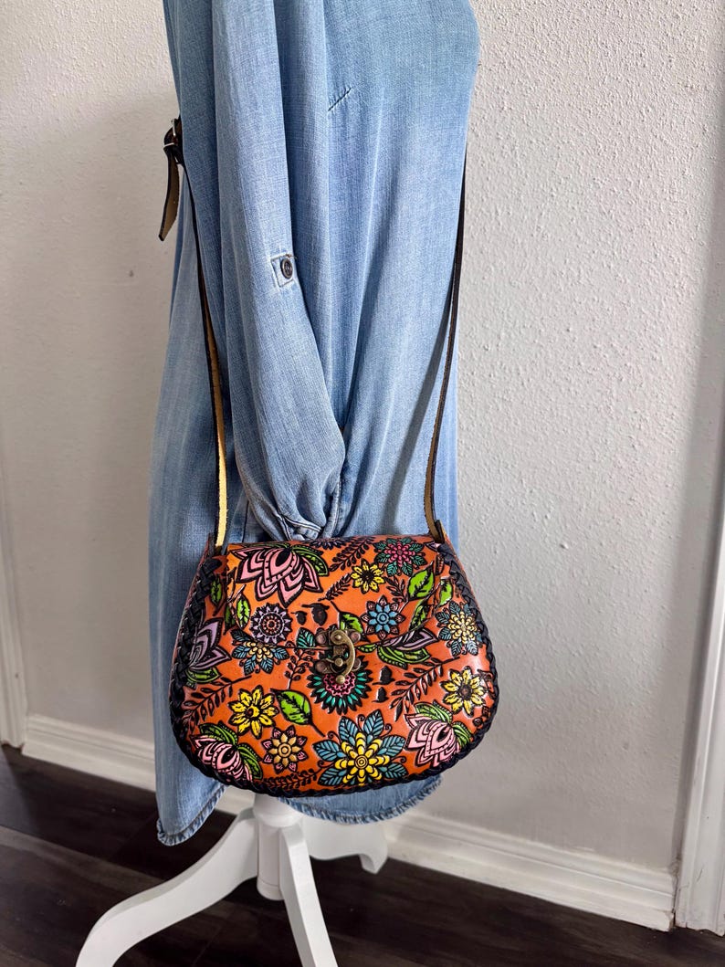 May include: Orange leather crossbody bag with a floral design in pink, green, blue, and yellow. The bag has a brown strap and a metal clasp. The bag is approximately 23 cm wide and 18 cm tall.