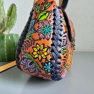 May include: A handcrafted leather shoulder bag with a vibrant floral design in orange, blue, yellow, and green. The bag features a braided navy blue trim and a black leather strap. The bag is a unique accessory.