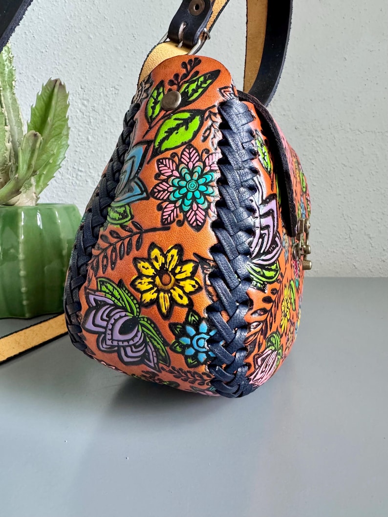 May include: A handcrafted leather shoulder bag with a vibrant floral design in orange, blue, yellow, and green. The bag features a braided navy blue trim and a black leather strap. The bag is a unique accessory.
