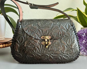 May include: Dark brown leather handbag with a floral rose pattern. The bag features a flap closure with a brass clasp, a braided edge, and a brown leather strap. The bag is designed with a shoulder strap and a top handle.