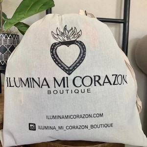 May include: A white canvas bag featuring a black heart and flame design, with the text "ILUMINA MI CORAZON BOUTIQUE". The bag also displays the website and Instagram handle.