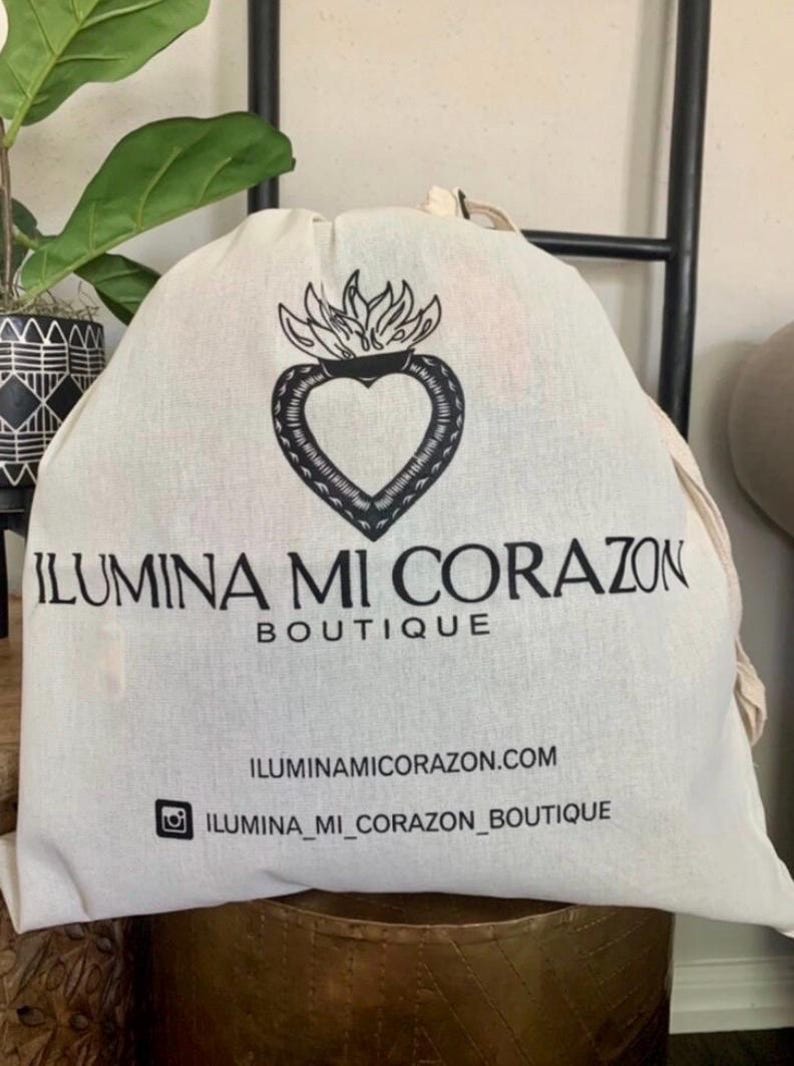 May include: A white canvas bag featuring a black heart and flame design, with the text "ILUMINA MI CORAZON BOUTIQUE". The bag also displays the website and Instagram handle.