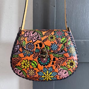 May include: Orange leather crossbody bag with a colourful floral design. The bag features a clasp closure and a woven black trim. The strap is a light tan colour. The floral pattern includes pink, yellow, blue, and green flowers.