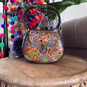 May include: An orange handbag with a floral pattern in pink, blue, yellow, and green. The bag has a black handle and a clasp closure. The bag is on a wooden table.