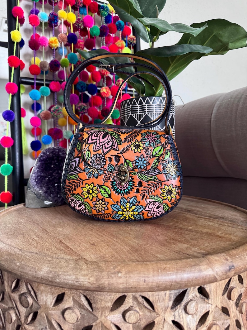 May include: An orange handbag with a floral pattern in pink, blue, yellow, and green. The bag has a black handle and a clasp closure. The bag is on a wooden table.