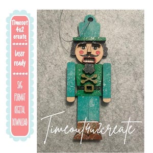 May include: A flat, wooden nutcracker ornament featuring a teal uniform with a distressed finish, a black belt with a gold buckle, and brown boots. The figure has a painted face with a mustache and a silver hanging string at the top.