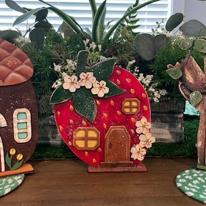 May include: Three miniature fairy houses made of wood. The first is shaped like an acorn with a brown roof and a red door. The second is shaped like a strawberry with a red body and a brown door. The third is shaped like a tree trunk with a brown bark and a blue door.