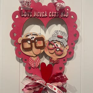 Op de afbeelding: Een houten wanddecoratie met Valentijnsthema. De rode hartvormige lijst toont een stel met brillen en de tekst "LOVE NEVER GETS OLD". Een rode strik en lint maken het ontwerp compleet.