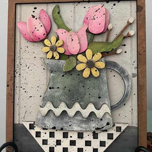 May include: A framed wooden wall art piece featuring a green pitcher with a white wavy rim, filled with pink tulips and yellow daisies. The pitcher sits on a black and white checkered plate.