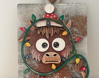 May include: A wooden Christmas ornament featuring a brown cow with large white eyes, wearing a red and white polka-dot hat. The cow is decorated with a green string of colorful Christmas lights. The ornament is mounted on a gray, weathered wooden plaque.