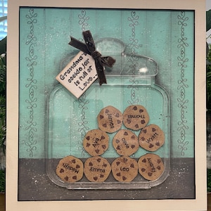 May include: A framed picture of a glass jar filled with cookies. The cookies are decorated with names, including Madilyn, James, Ava, Emma, Leo, Thomas, Audrey, Olivia, and Wyatt. The jar has a tag that reads "Grandma's cookie jar is full of love..."