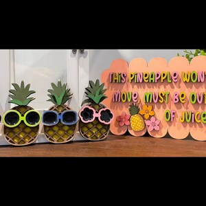 May include: Wooden pineapple decorations with sunglasses and a sign that reads "This Pineapple Won't Move, Must Be Out of Juice." The pineapples are yellow with green tops and various colored sunglasses.