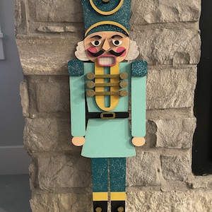 May include: A teal blue and gold wooden nutcracker figurine with a black hat, white beard, and a red mouth. The nutcracker is standing on a stone surface.