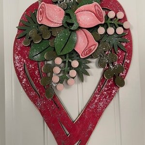 Könnte beinhalten: Rote, herzförmige Wanddekoration aus Holz mit floralem Arrangement. Die Anordnung umfasst rosa Rosen, grüne Blätter und rosa Beeren. Das Herz hat ein Used-Look-Finish mit silbernen Glitzerakzenten und wird von einer Schnurschlaufe gehalten.
