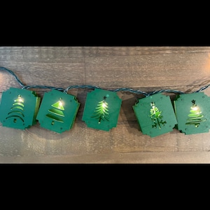 Peut inclure: Une guirlande de lumières de Noël vertes avec une découpe d'un sapin de Noël sur chaque lumière. Les lumières sont reliées entre elles par un fil transparent.