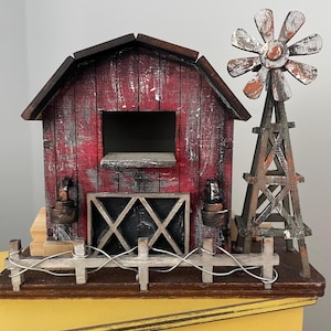 May include: A miniature wooden barn with a red and white painted exterior, a black and white gate, and a weathered windmill. The barn has a small window and a brown roof. The windmill is made of wood and has a white and brown painted design.