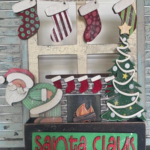 Op de afbeelding: Een houten kerstdecoratie met een kerstmanfiguur, een open haard met kousen, een kerstboom en een bord met de tekst "Santa Claus".