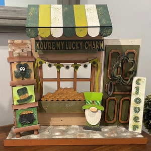 May include: A wooden St. Patrick's Day display with a green and white striped awning, a window with a pile of gold coins, and a leprechaun. The text "YOU'RE MY LUCKY CHARM" is above the window. Additional elements include a sign that says "CLOSED".