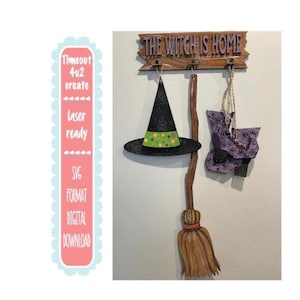 May include: A Halloween-themed wall hanging featuring a witch's hat, boots, and broom. The hat is black with a green and yellow band. The sign above reads "THE WITCH IS HOME."