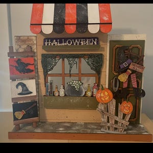 May include: A decorative Halloween shop display with a striped awning and the word "HALLOWEEN" above a window. The window features a cauldron, bottles, and books. A witch's hat and broom are near the door, and two carved pumpkins sit on a fence.