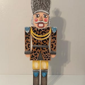 May include: A wooden nutcracker soldier with a leopard print jacket, yellow pants, and blue accents. The soldier has a stern expression, a white mustache, and a tall hat with a blue plume. The base is dark brown.