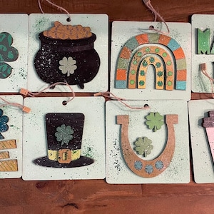 May include: Eight square wooden signs with a green, white, and brown paint splatter background. Each sign features a different St. Patrick's Day themed design, including a shamrock, pot of gold, rainbow, horseshoe, leprechaun hat, coffee cup, and the words "Love" and "March 17th".