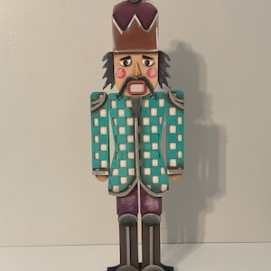 May include: A wooden nutcracker figurine with a teal and white checkered jacket, purple pants, and brown boots. The nutcracker has a brown hat with a purple top, a black mustache, and a red mouth. The figurine stands on a brown base.