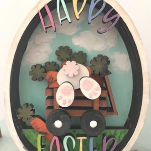 Fichier numérique svg Joyeuses Pâques oeuf glowforge. L'œuf est composé de plusieurs couches pour créer un effet 3D du lapin grimpant dans le chariot.