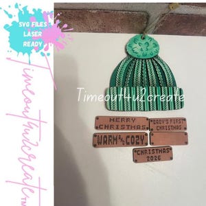 May include: A green wooden beanie ornament with a pom-pom and four rectangular wooden tags. The tags feature engraved text: MERRY CHRISTMAS, BABY'S FIRST CHRISTMAS, WARM AND COZY, and CHRISTMAS 2026. The text Timeout4u2create is visible.