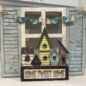 May include: A wooden sign with the text "Home Tweet Home" and a decorative window frame with blue bird cutouts and small birdhouses. The birdhouses are painted in shades of yellow, green, and brown.