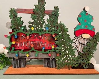 May include: A wooden Christmas gnome holding a red truck with a sign that says "Merry Christmas" and green pine trees. The truck is decorated with colorful lights.