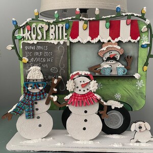 May include: A wooden Christmas-themed food truck with a green exterior and a red and white striped awning. The truck is decorated with white snow and a string of colorful lights. The truck says "Frost Bite" on the front and has a menu that reads "Snow Cones, Ice Cold Coco,  Avocado Ice, Ice Cream". There are three snowmen standing in front of the truck, one of which is holding a cup of hot cocoa. A white dog is standing in front of the truck's tire.