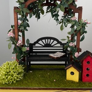 May include: Miniature garden scene featuring a black bench under a wooden arbor adorned with greenery and pink flowers. A small book rests on the bench. The scene includes a green bush and two colorful birdhouses, one yellow and one red, set on a grassy base.