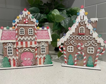 Pode incluir: Duas decorações de casas de gengibre com cobertura branca, detalhes de bengala de caramelo e granulado colorido. As casas são feitas de madeira e têm um acabamento brilhante.