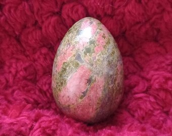 May include: A smooth, polished, green and pink egg-shaped stone with a marbled pattern. The stone is resting on a red fuzzy surface.