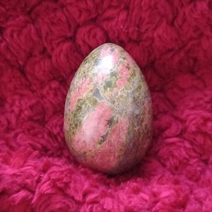 May include: A smooth, polished, green and pink egg-shaped stone with a marbled pattern. The stone is resting on a red fuzzy surface.