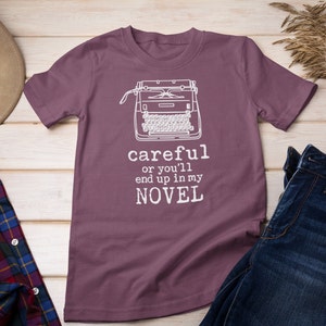 May include: A purple t-shirt with a white typewriter graphic and the text "careful or you'll end up in my NOVEL".