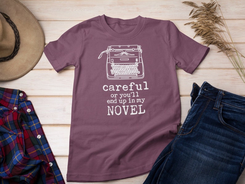 May include: A purple t-shirt with a white typewriter graphic and the text "careful or you'll end up in my NOVEL".