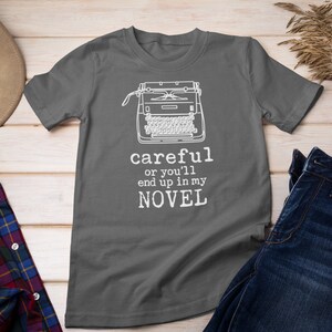 May include: A dark gray t-shirt with a white typewriter graphic and the text "careful or you'll end up in my NOVEL".