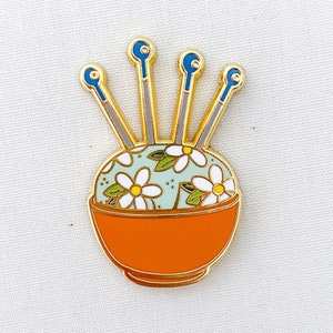 May include: Gold-tone enamel pin of a floral pincushion with four needles. The pincushion is in a small orange bowl with a white floral pattern.