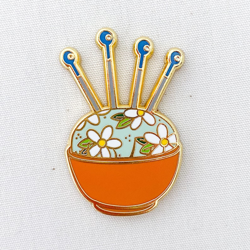 May include: Gold-tone enamel pin of a floral pincushion with four needles. The pincushion is in a small orange bowl with a white floral pattern.