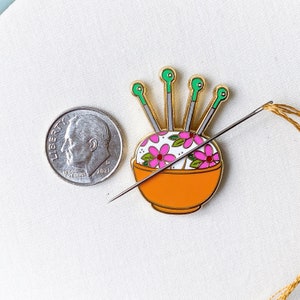 May include: Gold-tone pin of a floral pincushion with five green and gold needles sticking out of it. The pincushion is filled with pink flowers and has a white background.