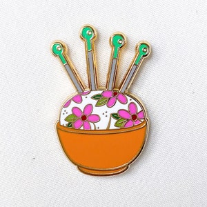May include: Gold pin featuring a floral pincushion and four green and gold needles. The pincushion is in a small orange bowl.